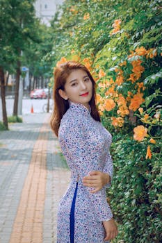 Asian woman in a floral Ao Dai posing beside vibrant orange flowers outdoors.